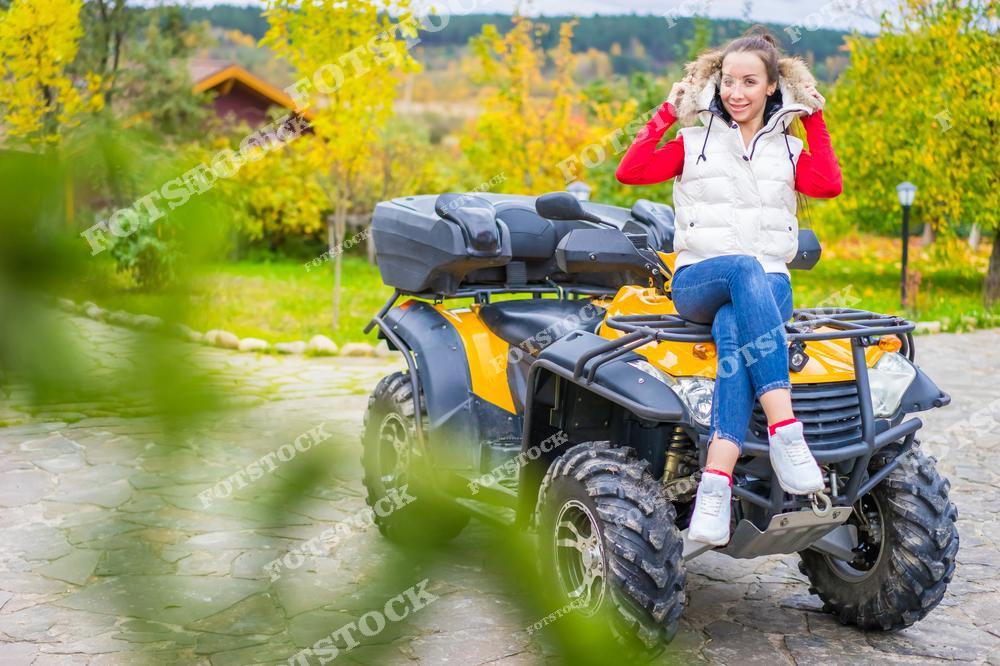 На фотографии изображена молодая женщина в теплой белой жилетке и красном свитере, сидящая на желтом квадроцикле. Она улыбается и держит капюшон куртки, создавая атмосферу радости и приключений. Фон представляет собой яркий осенний сад с желтыми и зелеными листьями.